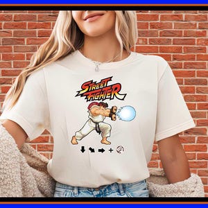 Puede incluir: Una camiseta de manga corta color crema con un gráfico del personaje Ryu de Street Fighter realizando un Hadouken. El diseño incluye el logotipo de Street Fighter, una esfera de energía azul y los símbolos de comando direccional debajo.