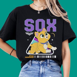 Peut inclure: Un t-shirt noir à manches courtes avec un motif représentant un chat orange nommé Sox. Le design comprend le texte SOX en grandes lettres violettes, un cadre triangulaire, une petite étoile et le texte STAR COMMAND avec un code-barres.