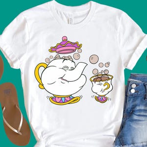 Puede incluir: Una camiseta blanca de manga corta con una ilustración gráfica de una tetera blanca con asa amarilla y tapa rosa, junto a una pequeña taza de té blanca con asa amarilla, ambas rodeadas de burbujas flotantes en la parte central frontal.