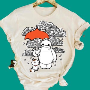May include: A cream-coloured short-sleeve t-shirt featuring a graphic print of a white robot holding a bright orange umbrella over a small cat. The background design consists of black and white line-art clouds and raindrops in a stylised pattern.