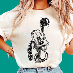 Może przedstawiać: Kremowy t-shirt z krótkim rękawem z czarnym szkicem postaci z kreskówki Pluto. Grafika przedstawia psa z jednym podniesionym uchem, szeroko otwartym pyskiem i wywieszonym długim językiem, wykonanym w luźnym, odręcznym stylu rysunkowym.