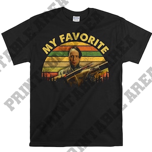 Puede incluir: Camiseta negra con un gráfico de estilo vintage de un hombre sosteniendo un rifle. El gráfico tiene un esquema de color rojo, amarillo y verde con el texto "My Favorite" arriba.