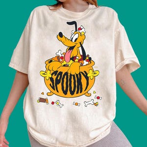 Może przedstawiać: Kremowy t-shirt z grafiką przedstawiającą żółtego psa siedzącego w pomarańczowym kociołku. Pies ma na głowie białą kokardę i wyciągnięty język. Słowo SPOOKY jest nadrukowane czarną czcionką na kociołku, w otoczeniu kości i cukierków.