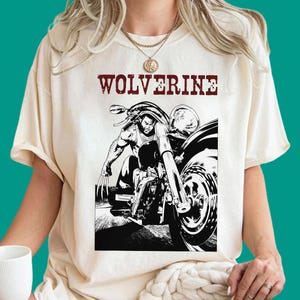 May include: A cream-coloured t-shirt featuring a black and white graphic print of a character with claws riding a motorcycle. The word WOLVERINE is printed in a distressed red font above the illustration. The shirt has a crew neckline and short sleeves.
