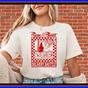 May include: A cream-colored short-sleeve t-shirt featuring a red graphic print. The design includes the text Pizza Planet, a rocket, a character, and three aliens inside a checkered border, with the text DELIVERY SHUTTLE SERVING YOUR LOCAL STAR CLUSTER.