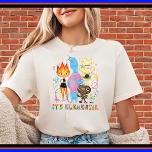 May include: A cream-coloured short-sleeve t-shirt featuring a colourful graphic of four animated characters from the movie Elemental. Below the characters, the text IT'S ELEMENTAL is printed in a multi-coloured, retro-style font with various small shapes.