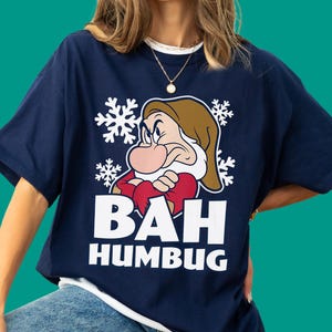 May include: A navy blue short-sleeve t-shirt featuring a graphic of the character Grumpy from Snow White wearing a brown hat, with white snowflakes in the background and the text BAH HUMBUG printed in large white capital letters below.