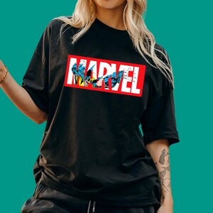 May include: A black short-sleeved t-shirt featuring a rectangular red graphic with the white text MARVEL. Inside the letters, there is a comic book illustration of the character Wolverine in a blue and yellow suit with extended metal claws.