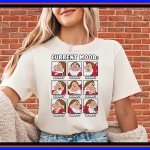 Peut inclure: Un t-shirt à manches courtes de couleur crème avec une impression graphique portant le texte "CURRENT MOOD:" au-dessus d'une grille de neuf carrés. Chaque carré affiche une illustration du personnage Grincheux avec le mot "GRUMPY" en dessous.