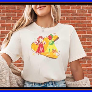 May include: A cream-coloured short-sleeve t-shirt featuring a graphic print of two cartoon characters, one in a red outfit and one in a green and yellow outfit, positioned over a yellow boomerang-shaped design with small star accents.