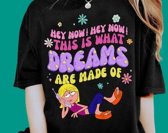 Pode incluir: Uma t-shirt preta com uma impressão gráfica com o texto "HEY NOW! HEY NOW! THIS IS WHAT DREAMS ARE MADE OF" em rosa, roxo e amarelo. O design inclui flores coloridas e uma personagem de desenho animado loira a usar um telefone.