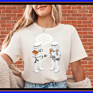 May include: A cream-coloured short-sleeve t-shirt featuring a graphic of two white stormtroopers. A speech bubble above them contains the text: Maybe these were the droids we were looking for. The stormtroopers are depicted in white armour with orange accents.