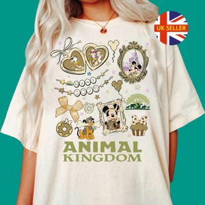 Może przedstawiać: Kremowy t-shirt z nadrukiem graficznym przedstawiającym postacie Disneya, w tym Myszkę Miki, Pluto i Kaczora Donalda. Wzór zawiera medalion w kształcie serca, ramkę z zamkiem, baner z napisem Name 2025 oraz słowa ANIMAL KINGDOM.