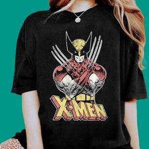 May include: A black short-sleeved t-shirt featuring a graphic print of the Marvel character Wolverine with his signature claws extended. Below the character, the text X-MEN is printed in a bold, yellow and red block-letter font with a distressed finish.