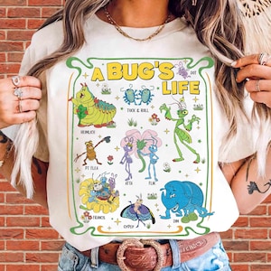 May include: A cream-coloured t-shirt featuring a colourful graphic print with the text A BUG'S LIFE. The design displays various animated insect characters including Heimlich, Tuck and Roll, P.T. Flea, Atta, Flik, Francis, Gypsy, and Dim, surrounded by small flowers and stars.