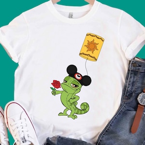 Puede incluir: Una camiseta blanca con un gráfico de un camaleón verde que lleva orejas de ratón negras con un emblema rojo en el centro. El camaleón sostiene una rosa roja y está atado a una linterna flotante amarilla con un diseño de sol al frente.