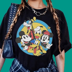 Może przedstawiać: Czarny t-shirt z krótkim rękawem z okrągłym nadrukiem graficznym przedstawiającym Myszkę Miki, Myszkę Minnie, Kaczora Donalda, Goofy'ego i Pluto. Postacie umieszczono w żółtym kole z niebieską obwódką, całość ma postarzany wygląd.