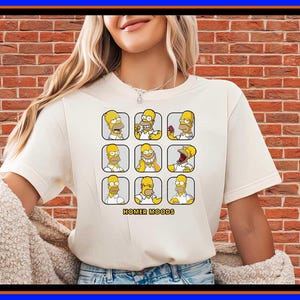 Pode incluir: Uma t-shirt de manga curta na cor creme com uma estampa gráfica de nove expressões faciais diferentes da personagem Homer Simpson dispostas numa grelha. Abaixo da grelha, o texto diz HOMER MOODS em letras maiúsculas amarelas.