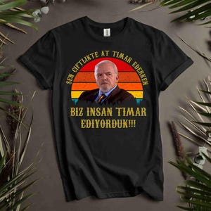 May include: Black t-shirt featuring a portrait of a man in a suit set against a sunset design. The shirt has text in a circular pattern around the portrait and below it. The shirt is displayed on a dark surface with green foliage.