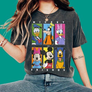 Może przedstawiać: Ciemnoszary t-shirt z krótkim rękawem z nadrukiem graficznym przedstawiającym sześć postaci Disneya w kolorowych prostokątnych ramkach. Napis MICKEY AND FRIENDS jest nadrukowany na biało nad i pod siatką postaci, w tym Kaczora Donalda, Goofy'ego, Daisy, Minnie, Myszki Miki i Pluto.