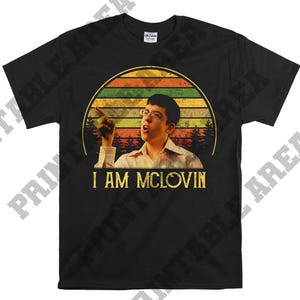 May include: Black t-shirt featuring a graphic of a man with glasses pointing, set against a sunset design. The text "I AM MCLOVIN" is printed in gold below the image. The shirt is made of a soft material.
