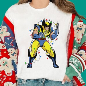 May include: A white t-shirt featuring a graphic of the comic book character Wolverine in a yellow and blue suit, with metallic claws extended, wrapped in a string of colourful Christmas lights, worn under a red cardigan with gingerbread men.