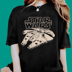Könnte beinhalten: Ein schwarzes Kurzarm-T-Shirt mit einer cremefarbenen Grafik des Millennium Falcon Raumschiffs aus Star Wars. Das ikonische Star Wars-Logo ist in Cremefarbe über der Illustration des Raumschiffs auf der Brust des Kleidungsstücks aufgedruckt.
