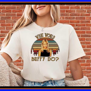 May include: A cream-coloured short-sleeve t-shirt featuring a graphic print of a blonde woman in a black top. The design includes a retro-style sunset background with trees and the text "WHAT WOULD BUFFY DO?" in a brown, arched, serif font.