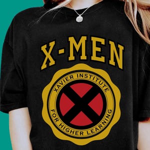 May include: A black short-sleeved t-shirt featuring the yellow text X-MEN above a circular emblem. The emblem contains the text XAVIER INSTITUTE FOR HIGHER LEARNING in yellow around a central red circle with a black X shape inside the circle.