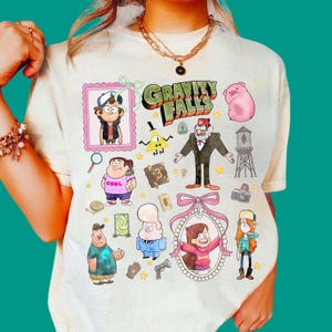May include: A cream-coloured t-shirt featuring a colourful graphic print of Gravity Falls characters including Dipper, Mabel, Grunkle Stan, Bill Cipher, and Waddles the pig, along with various show-related icons like a journal, camera, and water tower.