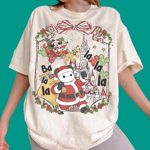 Może przedstawiać: Kremowy t-shirt z grafiką przedstawiającą Baymaxa przebranego za Świętego Mikołaja, Myszkę Miki i Minnie w saniach oraz Pluto. Wzór zawiera zamek, ostrokrzew, czerwono-białą kokardę oraz napis Ba la la la la la.