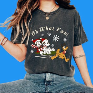 Może przedstawiać: Ciemnoszary t-shirt z krótkim rękawem z grafiką przedstawiającą Myszkę Miki i Minnie w czapkach Mikołaja na zielonych saniach ciągniętych przez Pluto. Nad postaciami znajduje się napis "Oh What Fun!" w otoczeniu białych płatków śniegu.