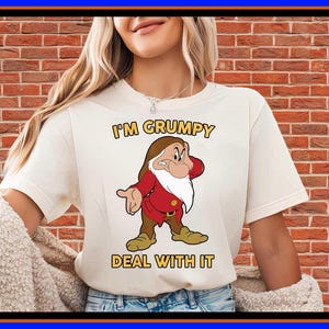 May include: A cream-coloured short-sleeve t-shirt featuring a graphic of the character Grumpy from Snow White. The text above the character reads I'M GRUMPY and the text below reads DEAL WITH IT in yellow block letters with a dark outline.