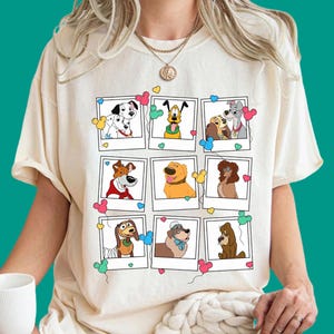 Może przedstawiać: Kremowy t-shirt z krótkim rękawem z nadrukiem graficznym przedstawiającym dziewięć pojedynczych ramek w stylu polaroid. Każda ramka zawiera inną postać psa z kreskówki, ozdobioną małymi kolorowymi balonami w kształcie serc i uszu myszy.