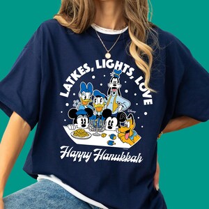 Może przedstawiać: Granatowy t-shirt z grafiką przedstawiającą postacie Disneya, w tym Myszkę Miki, Minnie, Kaczora Donalda, Daisy, Goofy'ego i Pluto świętujących Chanukę. Napis głosi "LATKES, LIGHTS, LOVE" oraz "Happy Hanukkah" z menorą i plackami latkes.