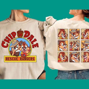 May include: A beige crewneck sweatshirt featuring a Chip 'n Dale Rescue Rangers graphic on the front with red text and an acorn icon. The back displays a grid of nine square panels showing various cartoon expressions of the two chipmunk characters.