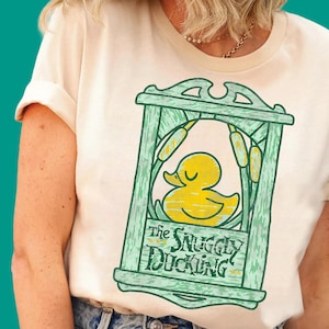 May include: A cream-coloured short-sleeve t-shirt featuring a graphic of a yellow rubber duck inside a green wooden-style frame. The design includes cattails and the text The Snuggly Duckling in a stylised dark green font on the lower section.