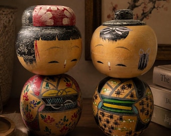 Könnte beinhalten: Zwei runde Kokeshi-Puppen aus Holz mit handbemalten Details. Die linke Puppe hat einen roten Blumenkopfschmuck und einen gelben Körper mit Blumenmustern. Die rechte Puppe hat einen schwarzen Haarknoten und einen gelben Körper mit schwarz-grünem Gittermuster.