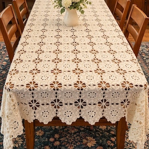 May include: A rectangular cream-colored crochet tablecloth featuring a repeating floral motif pattern with openwork detailing. The fabric drapes over the edges of a wooden dining table, showcasing intricate needlework and a scalloped border.