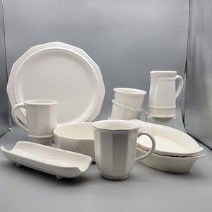 May include: A collection of white ceramic tableware, including a large octagonal plate, mugs, bowls, and serving dishes. The set features a classic, clean design, suitable for various dining occasions. The pieces are arranged on a light gray surface.