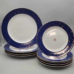 May include: A set of white ceramic plates with a navy blue border adorned with gold stars. The plates have a gold rim and are stacked in two groups, showcasing a celestial design. The plates are of different sizes.