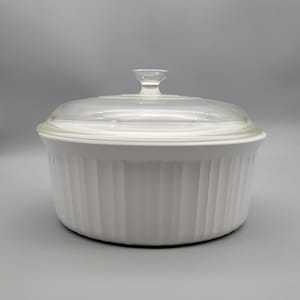 May include: White, round casserole dish with a clear glass lid. The dish has vertical ribbed detailing around the sides and a small handle on the lid. A classic kitchenware item.