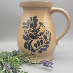 May include: A tan ceramic pitcher with a dark blue floral design. The pitcher has a curved handle and a spout. The floral design is on the front of the pitcher. The pitcher is sitting next to a sprig of lavender.