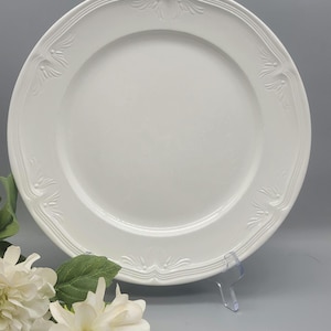 May include: A white ceramic dinner plate with a decorative embossed floral design around the rim. The plate is round and has a smooth, glossy surface. The plate is displayed on a clear plastic stand.