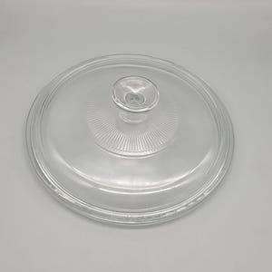 May include: A clear glass lid with a central knob and a ridged design. The lid is circular and appears to be for a pot or pan. The glass is transparent, allowing visibility of the contents below. The lid is likely for kitchen use.