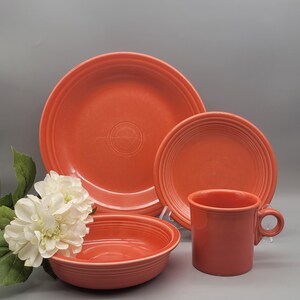 May include: A set of orange-red ceramic dinnerware, including a large plate, a smaller plate, a bowl, and a mug. The plates and bowl have concentric ring detailing. The mug has a handle. A white flower arrangement is in the background.