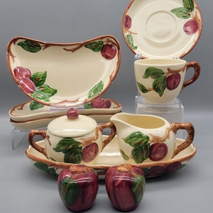 May include: A cream-colored ceramic dish set with a fruit design. The set includes a serving platter, a sugar bowl with a lid, a creamer, a cup and saucer, and two apple-shaped salt and pepper shakers. The fruit design features red apples and green leaves.