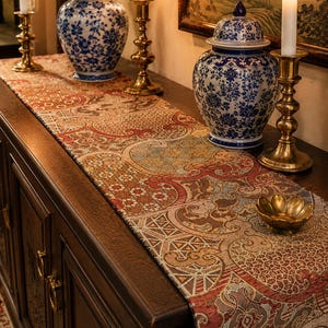May include: A long rectangular table runner featuring a complex woven pattern in shades of rust, gold, and blue. The fabric displays repeating geometric and floral motifs across its surface, laid flat on a dark wooden cabinet with brass hardware.