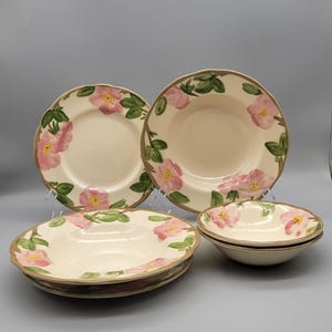 May include: A set of cream-colored ceramic plates and bowls. The plates and bowls feature a floral design with pink flowers, green leaves, and a brown rim. The set includes dinner plates, soup bowls, and smaller bowls.