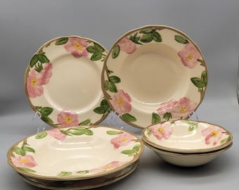May include: A set of cream-colored ceramic plates and bowls. The plates and bowls feature a floral design with pink flowers, green leaves, and a brown rim. The set includes dinner plates, soup bowls, and smaller bowls.